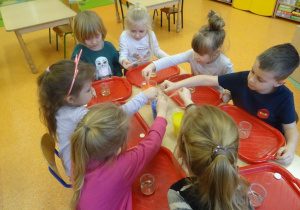 Dzieci nabierają pipetami zabarwione ciecze stojące w pojemnikach na środku stolika.